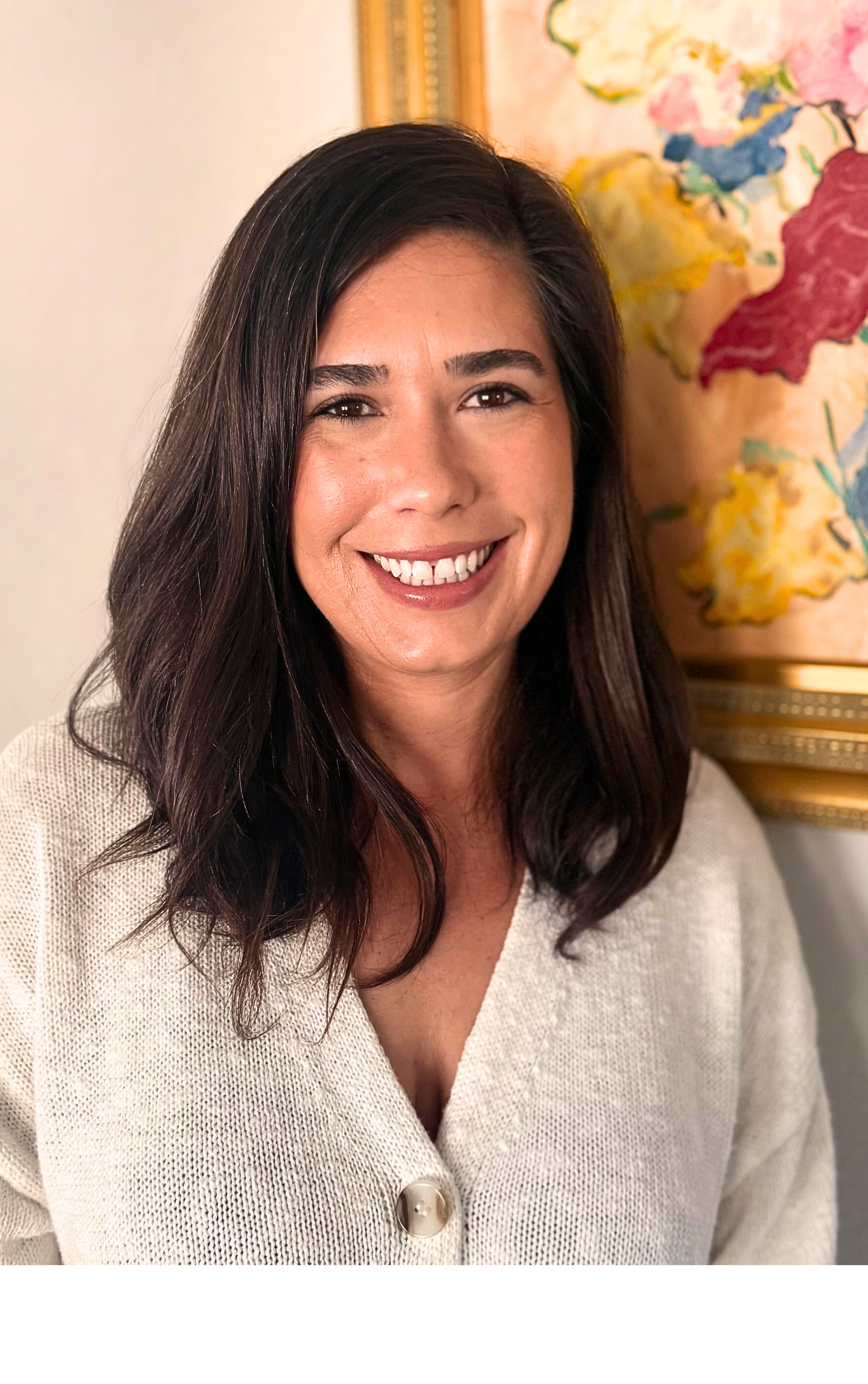 Woman with long dark hair wearing a light gray cardigan in front of a decorative wall piece.